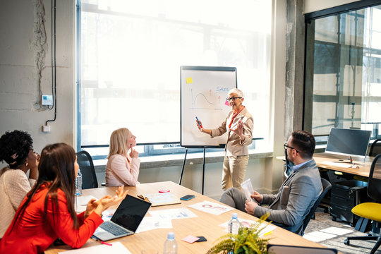 Aged attractive businesswoman giving presentation at corporate diverse group meeting, senior female business coach, woman boss or team leader presenting new strategic plan to multiracial employees - Powered by Adobe