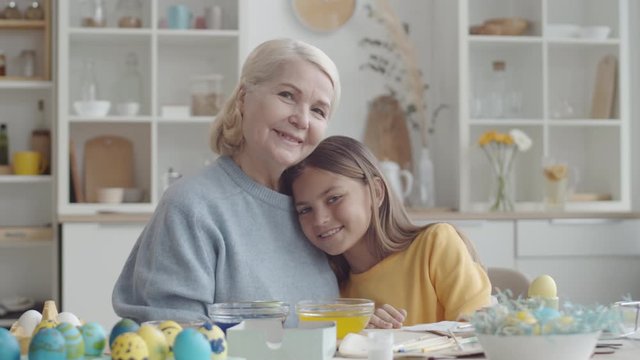 Lovely little girl and happy senior grandmother hugging each other, smiling and looking at camera while sitting together at kitchen table with Easter eggs on it