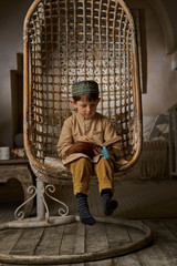 Obraz na płótnie Canvas Boy in arabic clothes with rosary beads reading holy quran book praying to Allah, prophet Muhammad holy spirit religion symbol concept inside eastern traditional interior