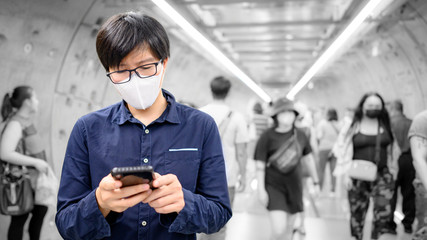 Asian man wearing surgical face mask using smartphone in subway tunnel with crowded people walking. Wuhan coronavirus (COVID-19) outbreak prevention in public area. Health care and medical concept