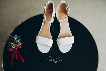 on a black table are white women's shoes, next to them are wedding rings and a boutonniere of burgundy flowers and greenery