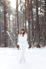 Fototapeta na wymiar A beautiful pretty young girl with curly hair and a snow-white smile in a white warm fur coat is walking and fooling around in the winter forest against the background of snow and trees, enjoying