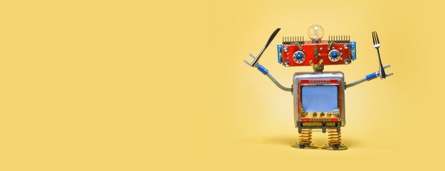 Hungry smiley robot holds fork and knife. Steampunk style robotic toy, blue monitor screen with copy space for text. Yellow background