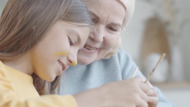 Cheerful little girl with yellow gouache on nose and cheek smiling and chatting with joyous grandmother while painting Easter egg together at table