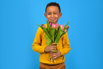 Portrait of a smiling African American boy with a bouquet of tulips for mom spring flowers on a blue background. Concept of greeting mother's Day, father's day, Valentine's day gift for a woman.