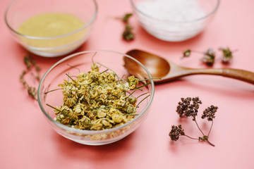 A natural skin care recipe. Dried chamomile in a glass bowl on a pink background of sea salt and yellow clay, natural powder. Create soothing and moisturizing cosmetics in a mortar at home