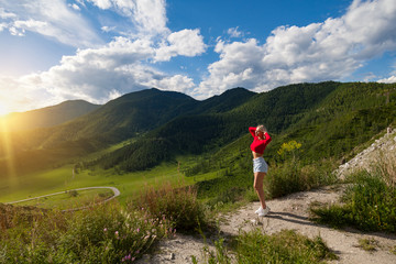 Fototapeta na wymiar The girl in red top and blue shorts reaches for the sun on the edge of a cliff in the Altai Mountains, below are green fields with trees and grass under a blue sky. Travel and vacation.