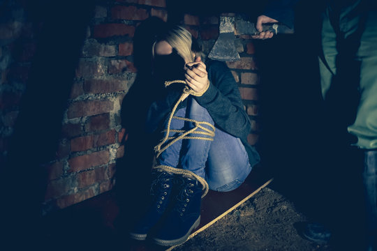irl with her hands and feet tied is sitting in the basement and screaming, and a man in rubber boots is standing next to her with an axe in his hands. Concept of violence, kidnapping