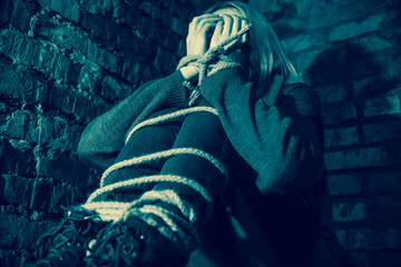 blonde girl with bound hands and feet is sitting in the basement, covering her face with her hands...