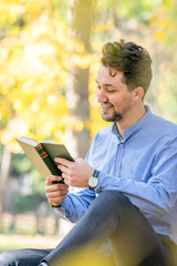 Fototapeta premium Handsome young man reading a book in a park. Portrait of a young man with a denim jacket and blue shirt reading a book outside. A guy reading a book in a park