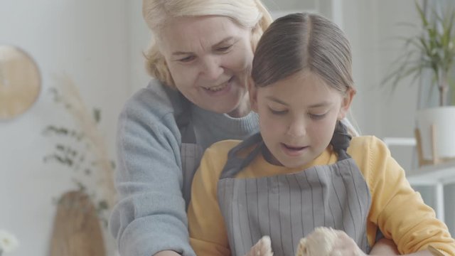 Handheld camera shot of eleven years old girl and her senior grandmother enjoying kneading dough together while preparing for baking Easter cookies