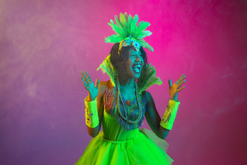 Happiness. Beautiful young woman in carnival, stylish masquerade costume with feathers dancing on gradient background in neon. Concept of holidays celebration, festive time, dance, party, having fun.