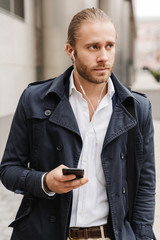 Image of young businessman walking on city street with cellphone