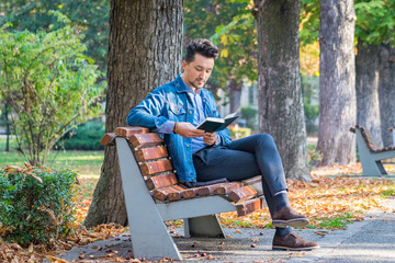 Handsome young man reading a book in a park. Portrait of a young man with a denim jacket and blue shirt reading a book outside. A guy reading a book in a park
