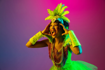 Astonished. Beautiful young woman in carnival, stylish masquerade costume with feathers dancing on gradient background in neon. Concept of holidays celebration, festive time, dance, party, having fun.