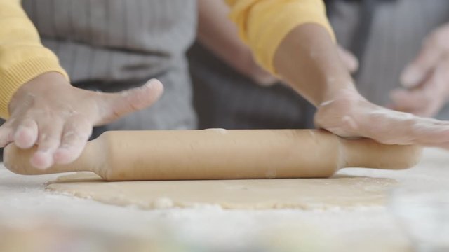 Close up shot of hands of little girl in apron rolling out pastry dough with wooden pin on kitchen table while baking with grandma at home