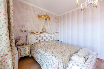 Fototapeta na wymiar Elegant Royal white bedroom with a large double bed. White Carved back with a small canopy, white bedside tables with curved legs. Light striped Wallpaper. Lamps and chandelier 