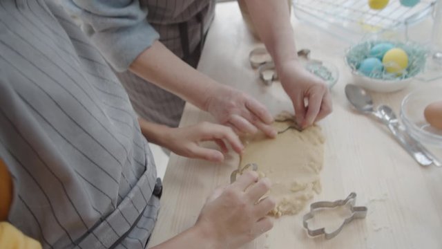 Tilt down shot of little girl and her grandmother cutting dough into rabbit forms and putting cookies on round baking sheet while getting ready for Easter holiday
