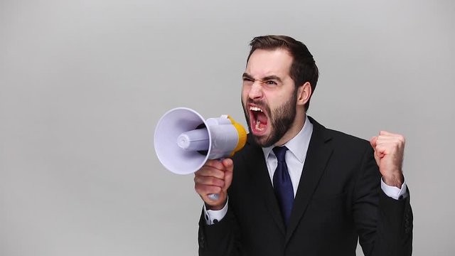 Angry mad bearded young business man in classic black suit shirt posing isolated on grey background studio. Achievement career wealth business concept. Mock up copy space. Scream in megaphone Hurry up