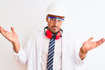 Young chemist man wearing security helmet and headphones over isolated background clueless and confused expression with arms and hands raised. Doubt concept.