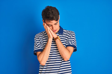 Teenager boy wearing casual t-shirt standing over blue isolated background sleeping tired dreaming and posing with hands together while smiling with closed eyes.