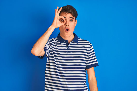 Teenager boy wearing casual t-shirt standing over blue isolated background doing ok gesture shocked with surprised face, eye looking through fingers. Unbelieving expression.