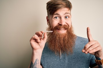 Irish redhead man with beard holding dental invisible aligner for tooth correction surprised with an idea or question pointing finger with happy face, number one