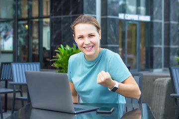 Happy cheerful successful girl with laptop in cafe outdoors, smartphone, young beautiful woman smiling, looking at camera, celebrating success, victory, good news. Got a job, passed an interview