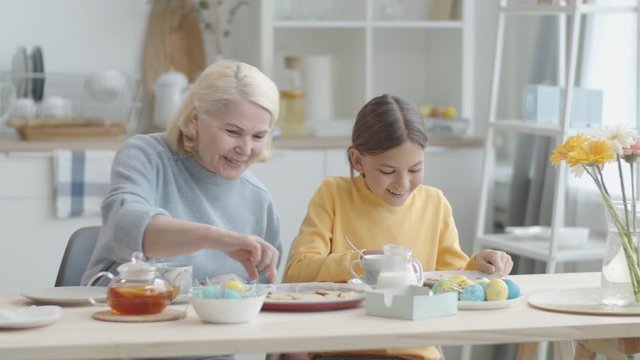 Happy grandmother smiling and talking while bringing just baked Easter cookies and pouring tea into cup for little granddaughter at kitchen table