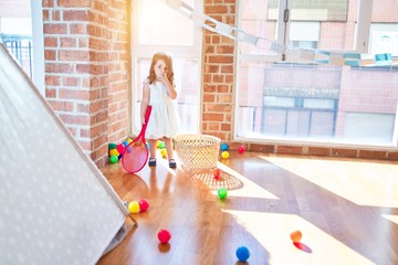 Adorable blonde toddler playing tennis using racket and ball around lots of toys at kindergarten