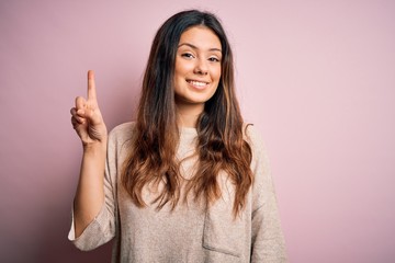 Young beautiful brunette woman wearing casual sweater standing over pink background showing and pointing up with finger number one while smiling confident and happy.