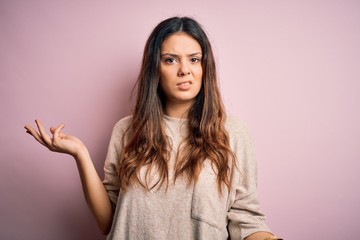 Young beautiful brunette woman wearing casual sweater standing over pink background clueless and...