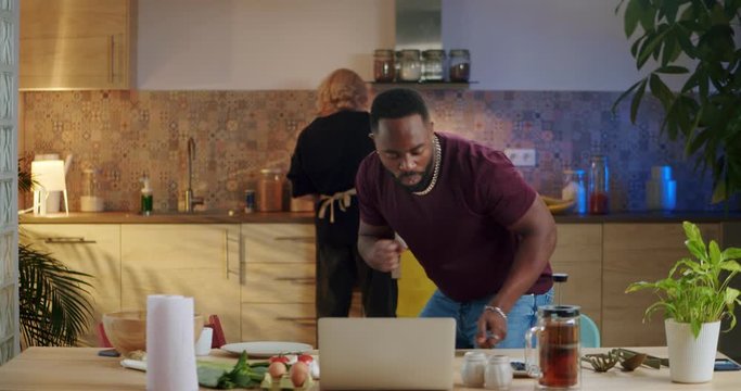 Affectionate mixed race couple joyful morning time together at kitchen leisure lifestyle, guy in good positive mood dancing serene while blonde woman cooking breakfast eggs early time indoor
