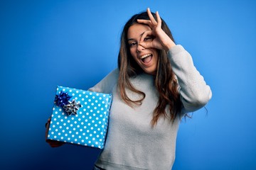 Young beautiful brunette woman holding birthday gift over isolated blue background with happy face smiling doing ok sign with hand on eye looking through fingers