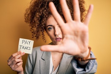 Young business woman with curly hair holding pay taxes to goverment reminder over yellow background...