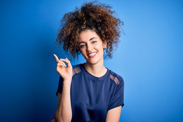Young beautiful woman with curly hair and piercing wearing casual blue t-shirt with a big smile on face, pointing with hand and finger to the side looking at the camera.