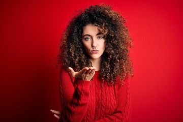 Young beautiful woman with curly hair and piercing wearing casual red sweater looking at the camera blowing a kiss with hand on air being lovely and sexy. Love expression.