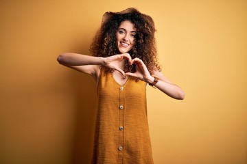 Beautiful brunette woman on vacation with curly hair and piercing wearing hat and dress smiling in love doing heart symbol shape with hands. Romantic concept.