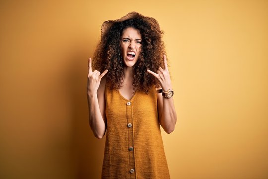 Beautiful brunette woman on vacation with curly hair and piercing wearing hat and dress shouting with crazy expression doing rock symbol with hands up. Music star. Heavy concept.
