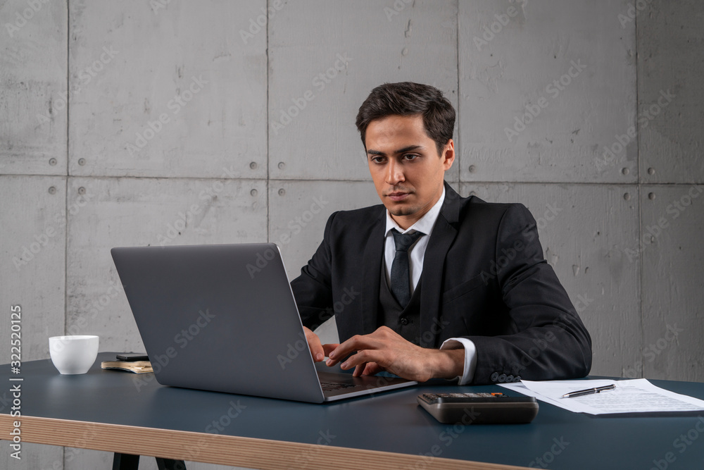 Wall mural portrait of serious and concentrated young man in dark suit typing on laptop in loft office. concept