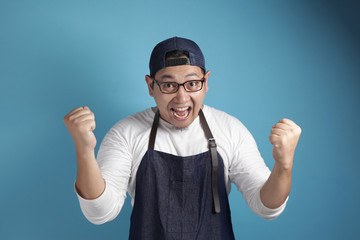 Portrait of happy excited proud Asian chef or waiter shows winning gesture, against blue background