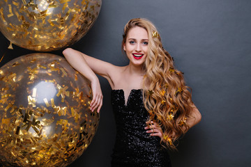Naklejka premium Portrait charming joyful young woman in black luxury dress, with long curly blonde hair, big balloons full with golden tinsels on black background. Celebrating birthday party, expressing positivity