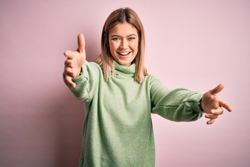 Young beautiful blonde woman wearing winter wool sweater over pink isolated background looking at the camera smiling with open arms for hug. Cheerful expression embracing happiness.