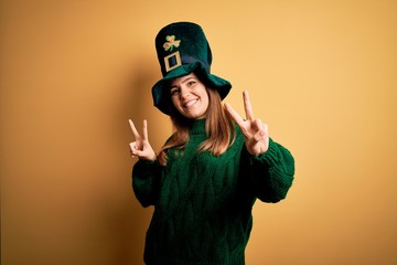 Young beautiful brunette woman wearing green hat on st patricks day celebration smiling looking to the camera showing fingers doing victory sign. Number two.