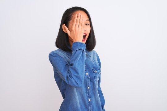 Young beautiful chinese woman wearing denim shirt standing over isolated white background covering one eye with hand, confident smile on face and surprise emotion.