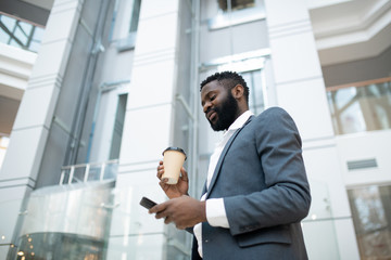 Below view of bearded Black businessman in jacket answering in messenger and drinking coffee on move