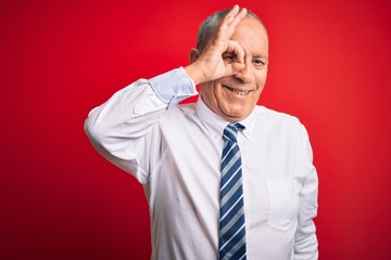 Senior handsome businessman wearing elegant tie standing over isolated red background doing ok gesture with hand smiling, eye looking through fingers with happy face.