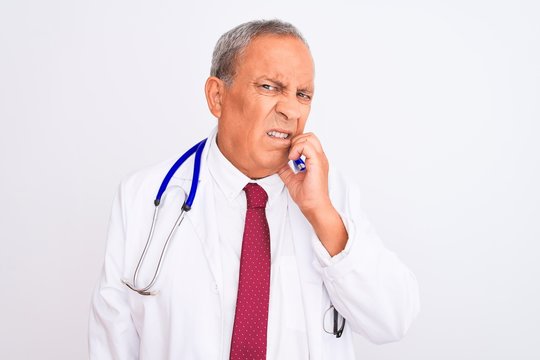 Senior grey-haired doctor man wearing stethoscope standing over isolated white background looking stressed and nervous with hands on mouth biting nails. Anxiety problem.