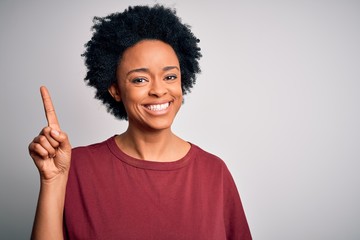 Young beautiful African American afro woman with curly hair wearing casual t-shirt standing showing and pointing up with finger number one while smiling confident and happy.