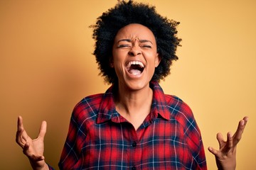 Young beautiful African American afro woman with curly hair wearing casual shirt crazy and mad shouting and yelling with aggressive expression and arms raised. Frustration concept.